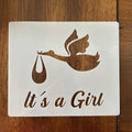 Decorative sign with stork carrying a baby and 'It's a Girl' text on a wooden surface