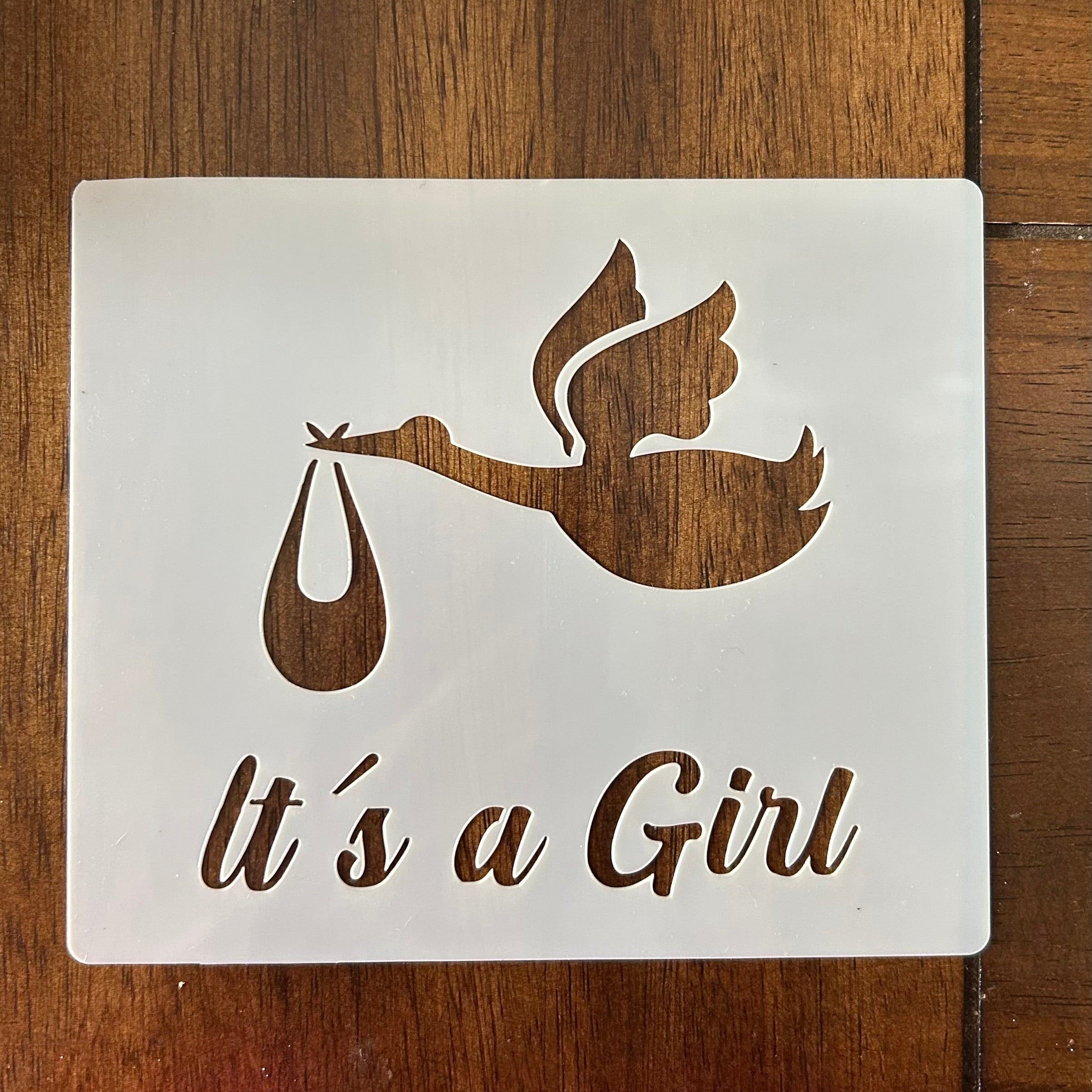 Decorative sign with stork carrying a baby and 'It's a Girl' text on a wooden surface